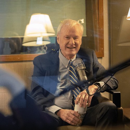 Chris Matthews sits in front of a microphone. He is smiling and holding a mug.