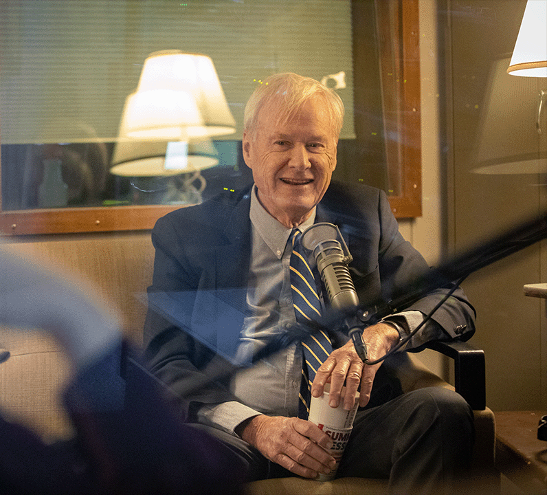 Chris Matthews sits in front of a microphone. He is smiling and holding a mug.