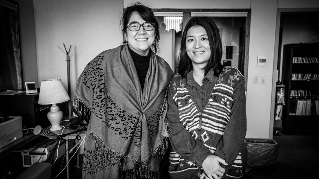 Sueann Ramella, left, and Jiemei Lin, right, stand side-by-side for a photo. Both are smiling. Jiemei Lin has her hands clasped in front of her, Sueann has hers behind her.