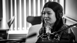 Jiemei Lin sits on a couch with headphones on her head, answering a question for Sueann Ramella. A microphone sits in front of Jiemei, partially obscuring her chin. The photo's background is out-of-focus.