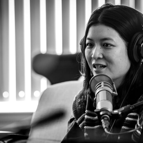 Jiemei Lin sits on a couch with headphones on her head, answering a question for Sueann Ramella. A microphone sits in front of Jiemei, partially obscuring her chin. The photo's background is out-of-focus.