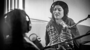 On the left side of the image, out of focus in the foreground, is the back of Sueann Ramella's head. On the right side of the photo is Jiemei Lin, who sits across from Sueann on a couch. She appears to sit on her hands and look toward the floor as she answers a question. They both wear headphones and have microphones in front of them.