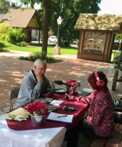 Jim Dee sits across a table from NWPB Producer Ash Beard. Jim is speaking into a microphone and Ash is listening through headphones. They are outside of the Dayton Depot Historical Museum.
