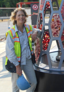 This is a photo of listener, Lin McJunkin. She is wearing a neon vest and leaning against a metal sculpture.