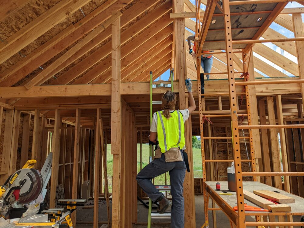 A woman in a bright green safety vest hands a tool to a woman in an orange safety vest as they work on an unfinished house.