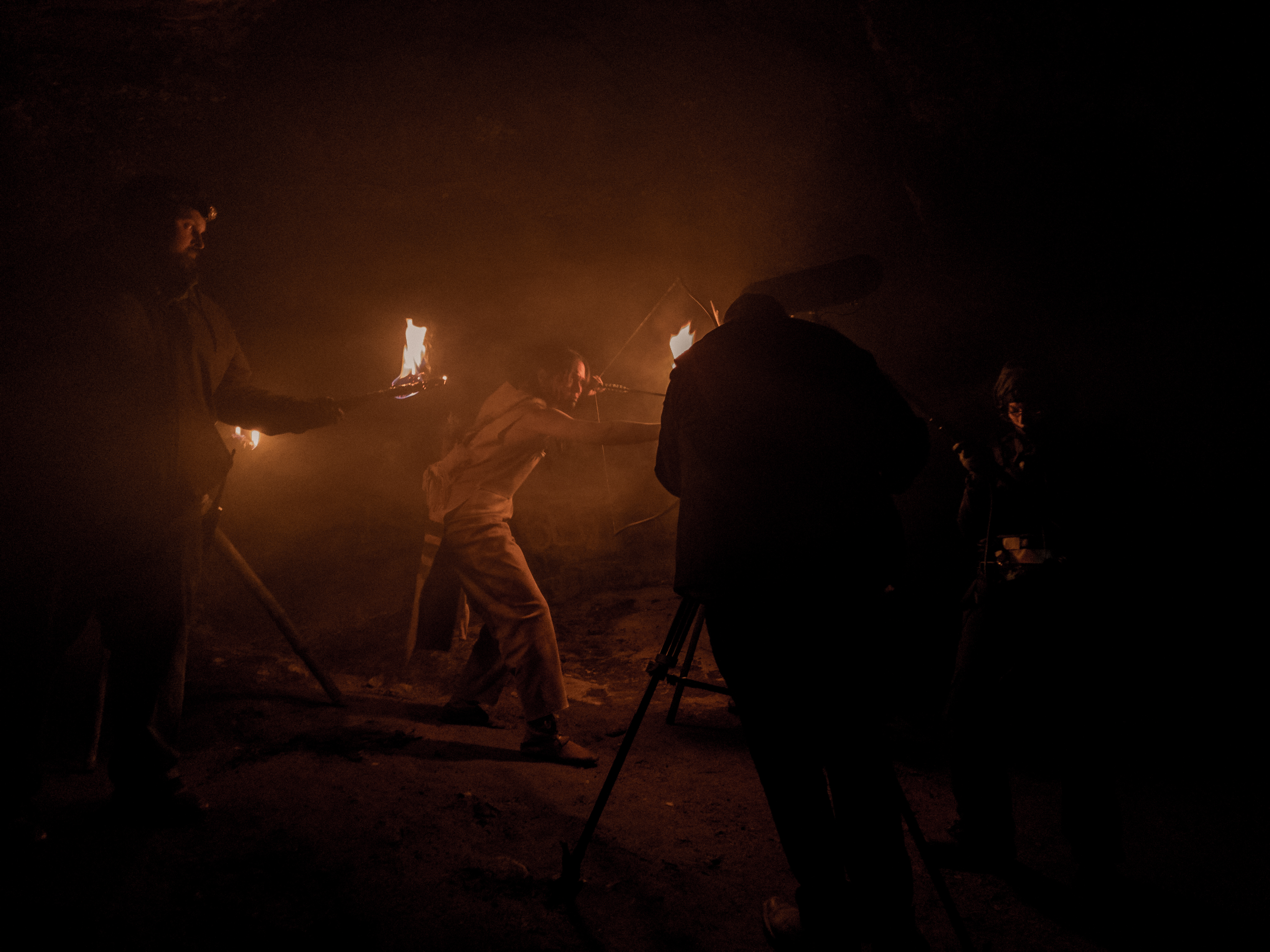 A nighttime scene shows an actor holding a bow and arrow in front of the camera, lit by torch light.