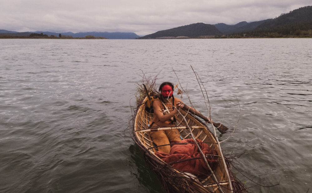 An actor in traditional Spokane tribal clothing canoes on the water of a river with stormy clouds overhead.