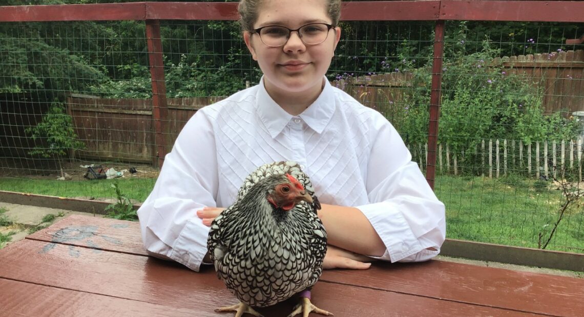 A young lady with glasses and her hair in a bun sits at a red picnic table with a white and black chicken on the table in front of her.