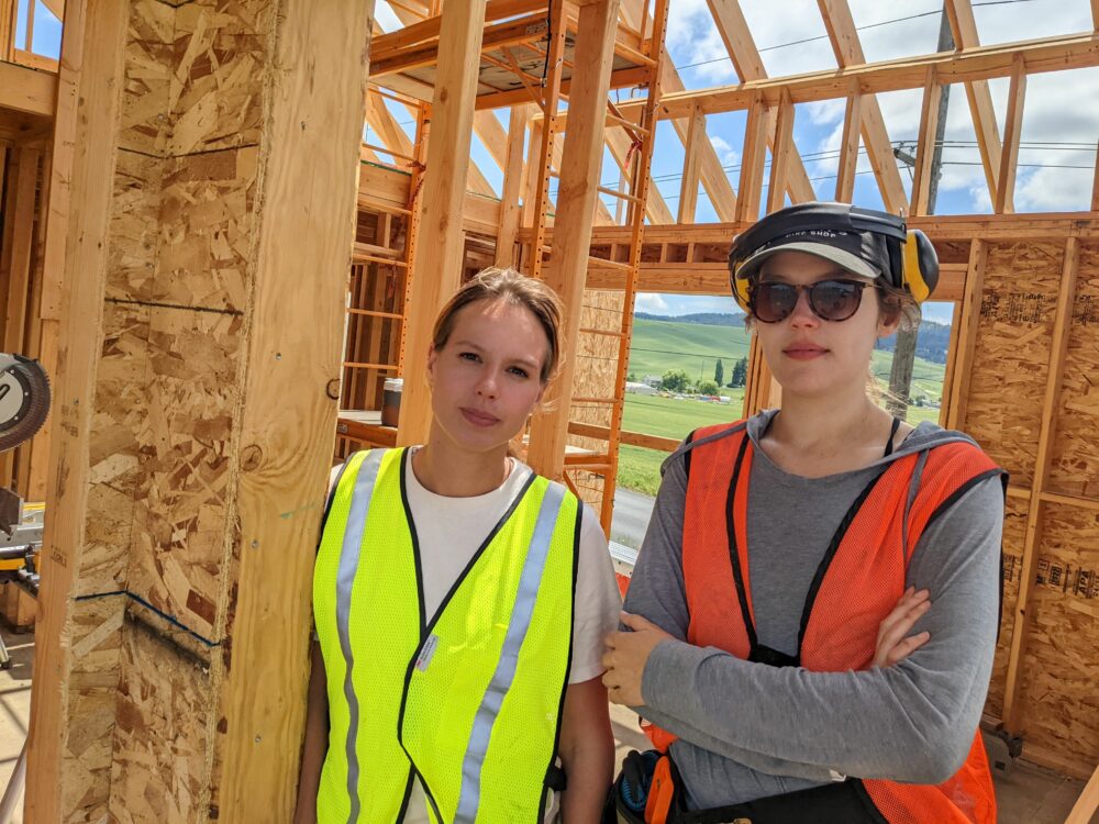 Two women in safety vests stand in the light brown frame of an unfinished house. 