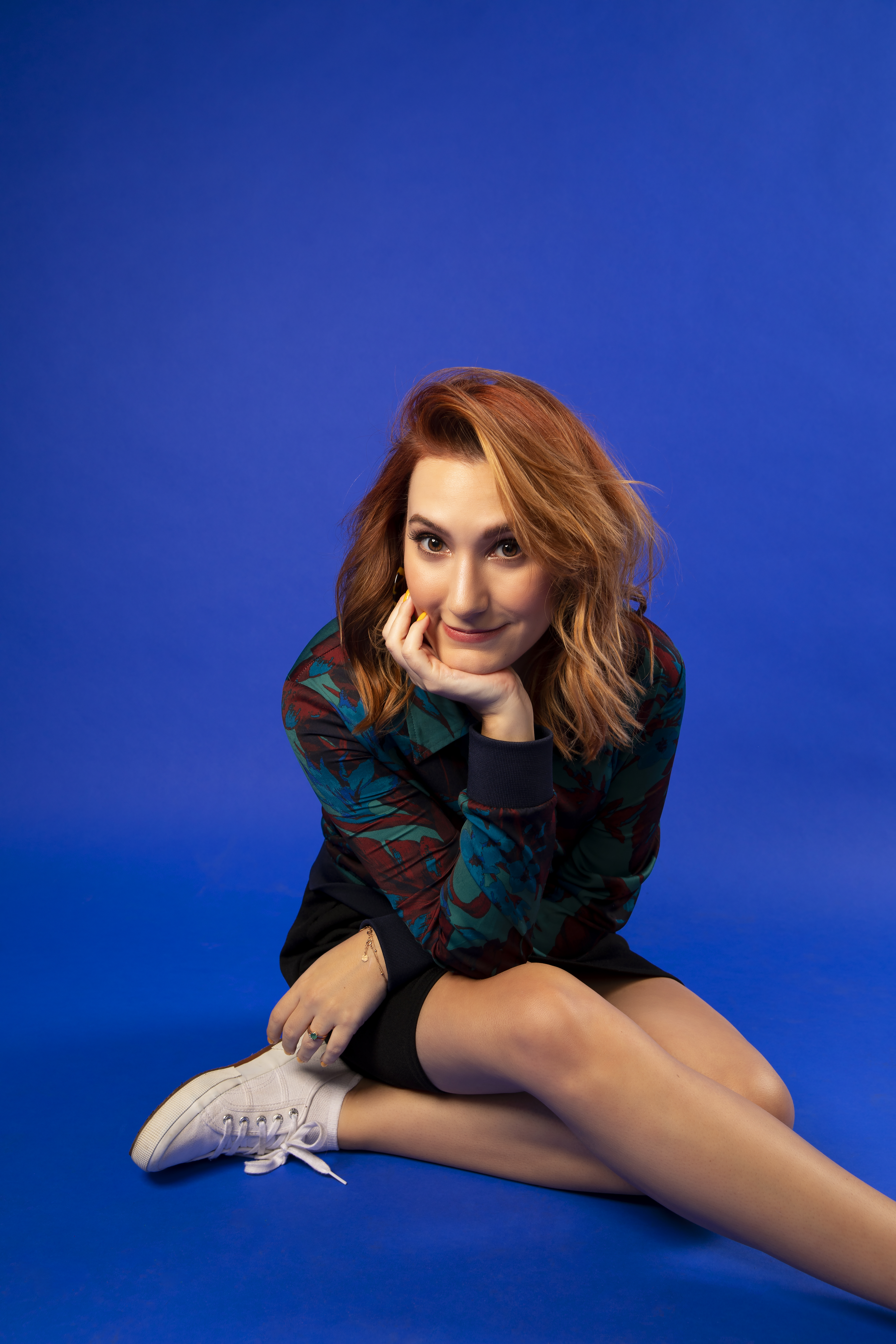 Sam Maggs sits in front of a royal blue background. She wears white tennis shoes and a patterned jackets. She has medium length red hair and she is sitting crosslegged with her head in her palm.