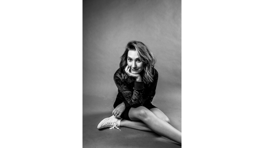 Studio photo of Sam Maggs. She sits on the ground, one leg crossed over the other and her head resting in her hand.