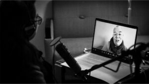 On the left side of the photo is Sueann, with glasses and headphones on and a microphone in front of her. On the right side of the photo, Petra is full-screen on a laptop, talking to Sueann.