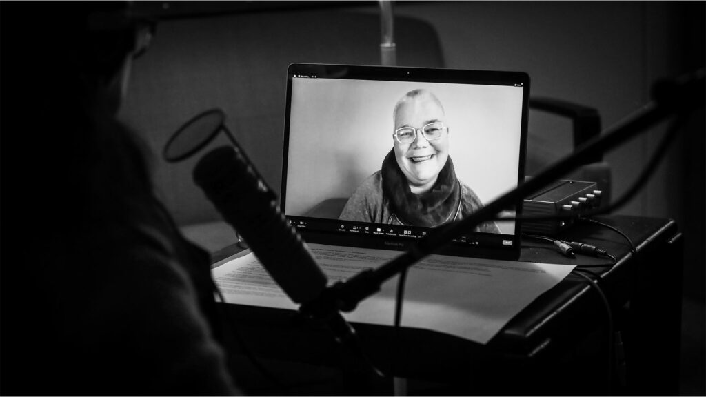 Petra, smiling, appears via Zoom on a laptop screen in the center of the photo. The back of Sueann's head and a microphone also appear, silhouetted from the laptop screen's light.