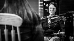 In this photo of Sarah Zabel she has her hands wide open in front of her as she looks across the roo to Sueann who sits with her back facing the camera. Sarah looks like she is mid-sentence at this moment.