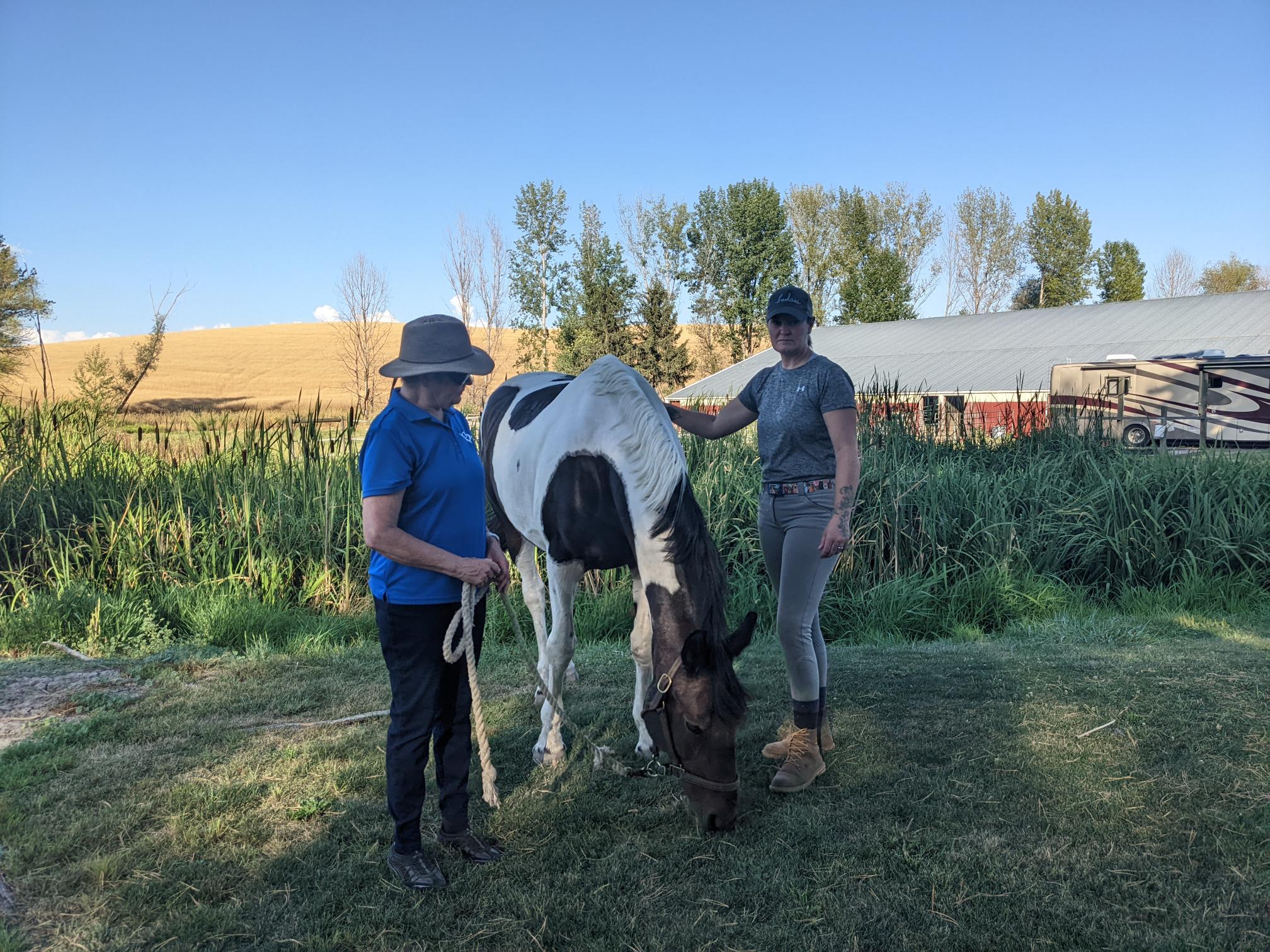 Dee and Erin Tomson stand near one of their horses on green grass next to a pond against a blue sky.