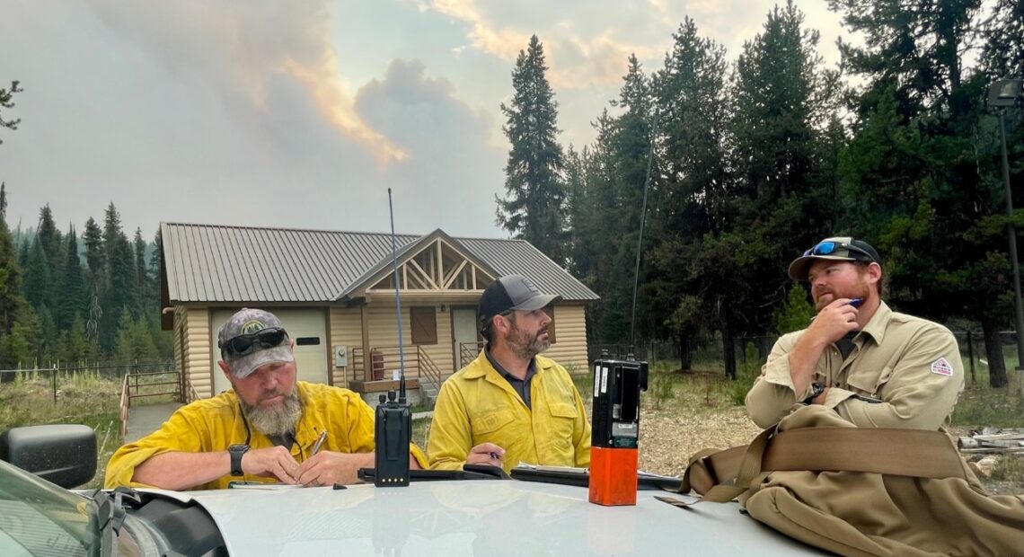 A group of men in tan and yellow shirts sit at a table in front of a log house near evergreen trees.