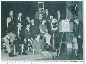 This is an old yearbook photo. Nancy Mack and two other women sit in a room full of men. Nancy sits on a stool near a large camera.