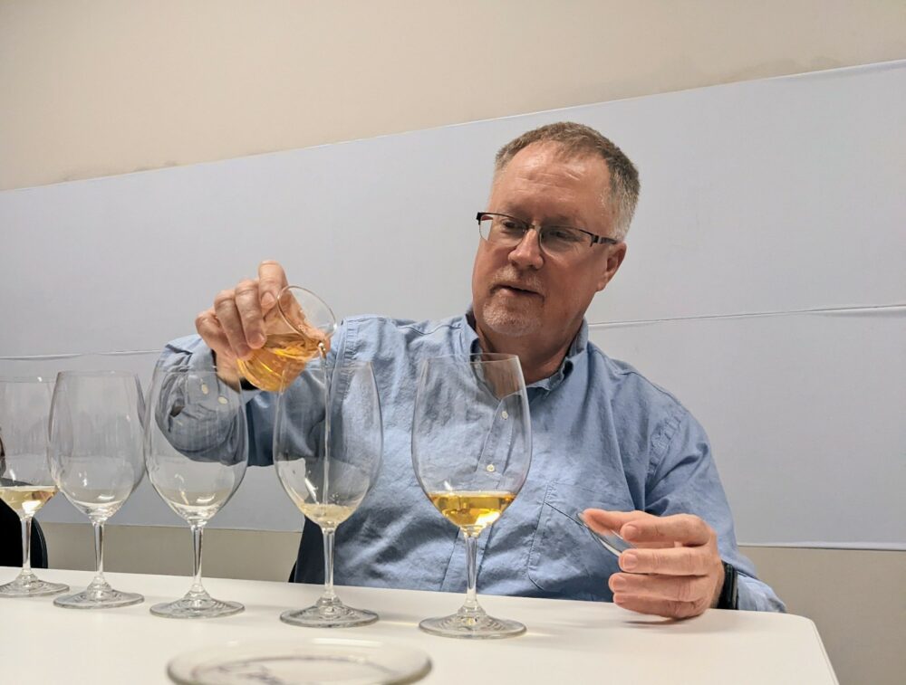 Tom Collins sits at a white table with a blue shirt on pouring yellow scotch into a wine glass. 