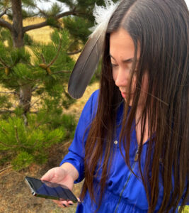A young Native American girl with long black hair and a bright blue shirt holds a mobile phone with the Camp Crier app.