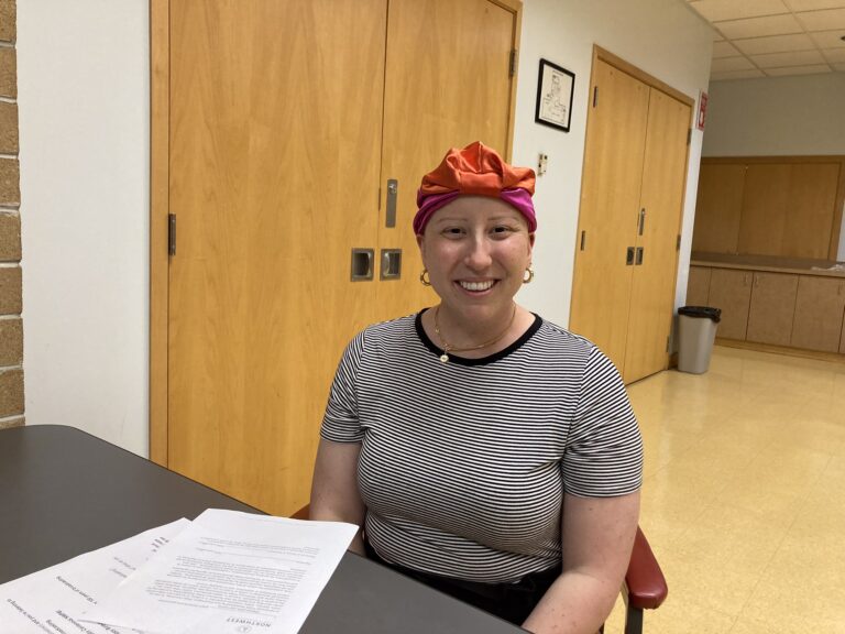 This is a photo of Madison. She sits at a table a nd smiles. She is wearing a head scarf and striped shirt.