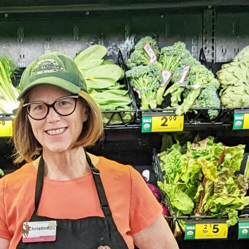 A women with red hair, black glasses, and orange shirt and a green hat poses in front of a produce section at Fred Meyer in Richland, Washington.