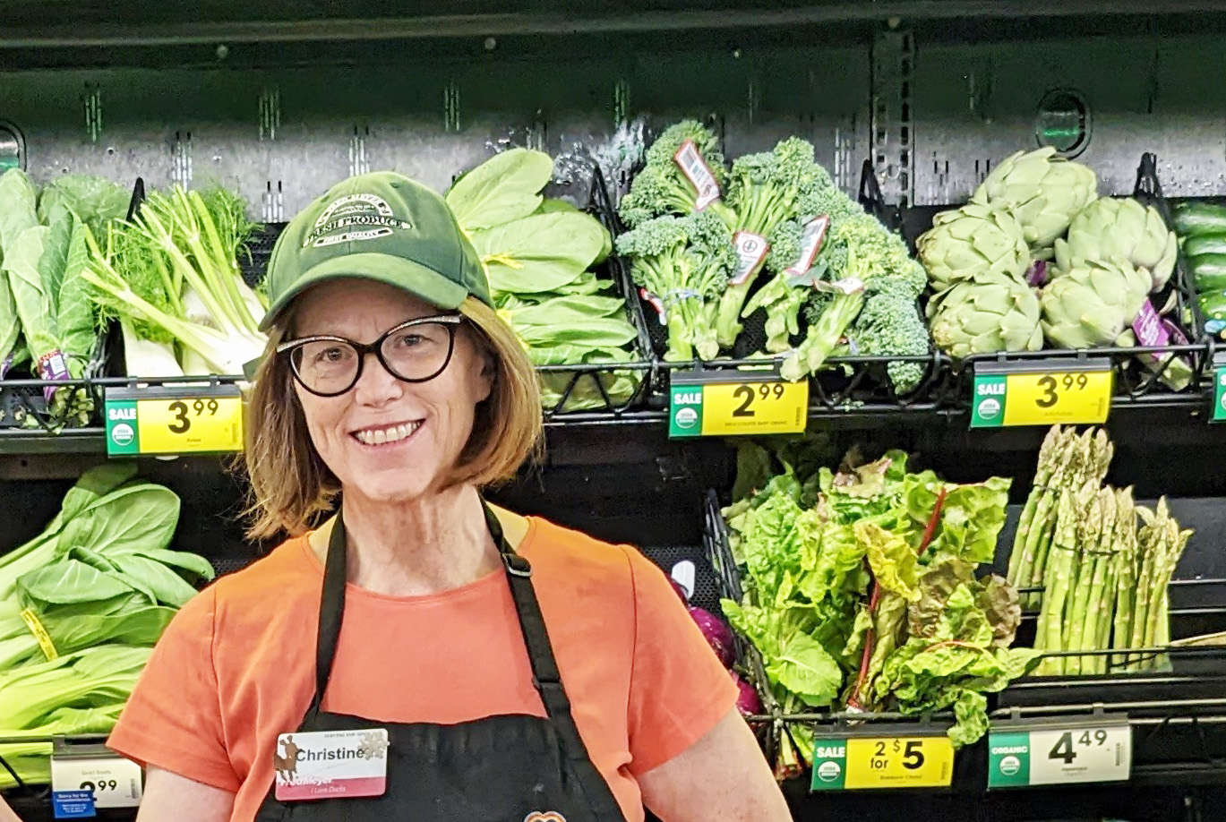 A women with red hair, black glasses, and orange shirt and a green hat poses in front of a produce section at Fred Meyer in Richland, Washington.