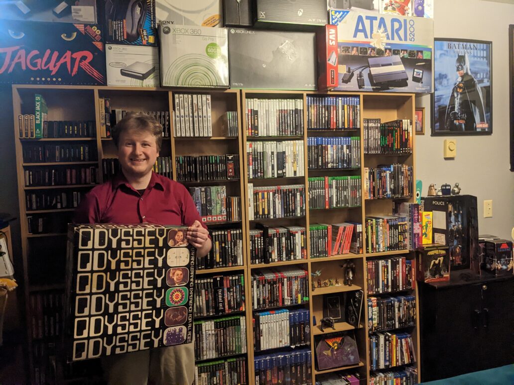 A blond guy in a red shirt holds an old game console in front of a bookshelf full of old video games.