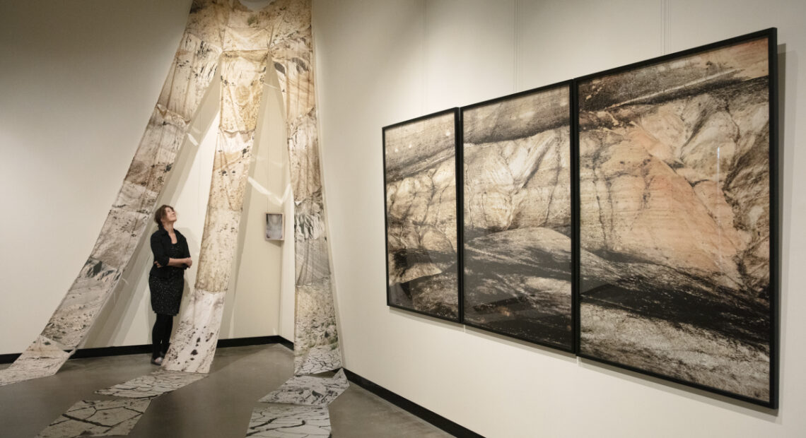 Artist Glenna stands in a black dress beneath photo collaged fabric draped from the ceiling near three framed photos of white bluffs.