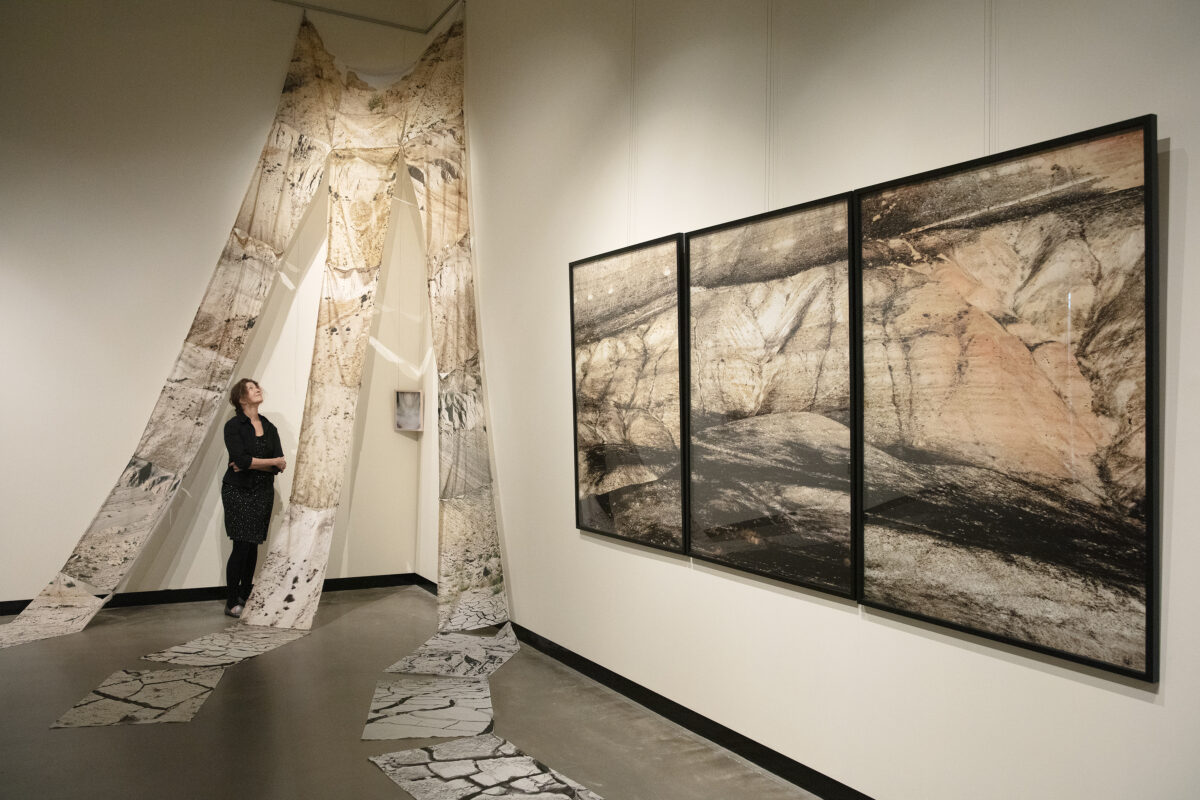 Artist Glenna stands in a black dress beneath photo collaged fabric draped from the ceiling near three framed photos of white bluffs.