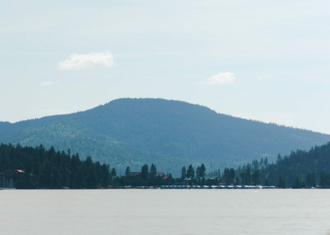A blue mountain rises beyond a dark green treeline along a blue lake.