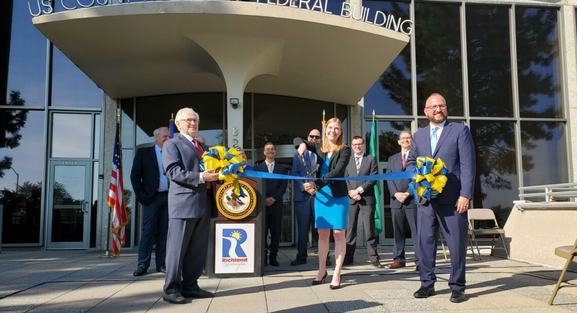 One older man, one woman, and one Hispanic man holding a ribbon in front of a builiding.