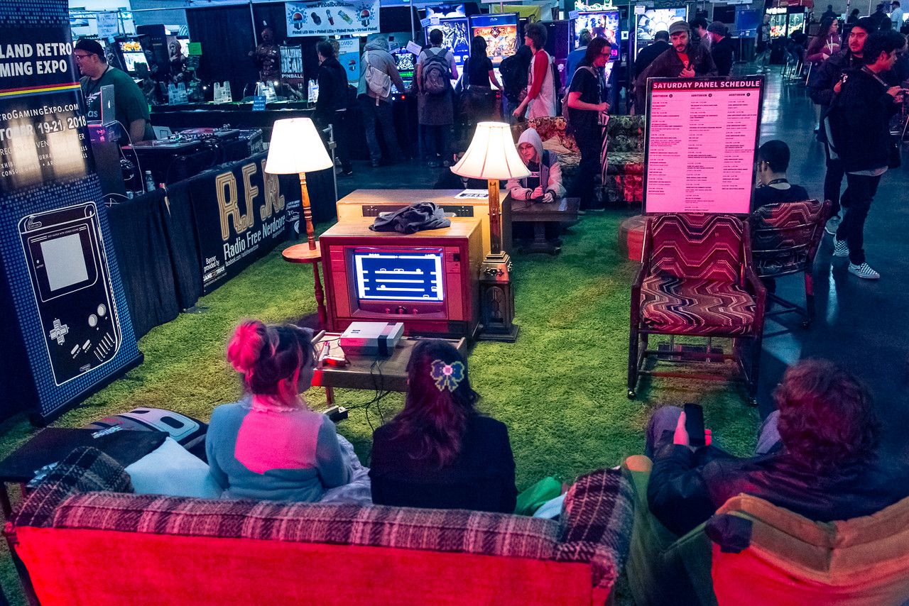 Two women with fun colors in their hair sit on a plaid couch in front of a TV surrounded by two lamps on a green carpet.