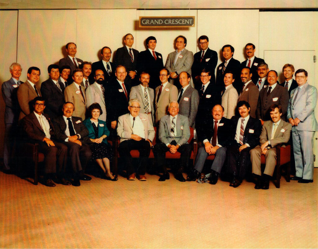 A reunion photo of around 30 people dressed up for an occasion in a half circle.