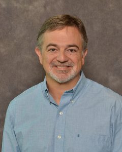 A man in a blue button down shirt with light brown hair and a gray beard.