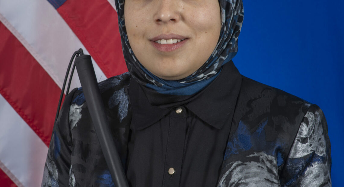 This is a photo of Sara Minkara. She wears a dark blue and black blazer with a patterned hijab. She holds a cane and stands in front of an American flag. She is smiling.