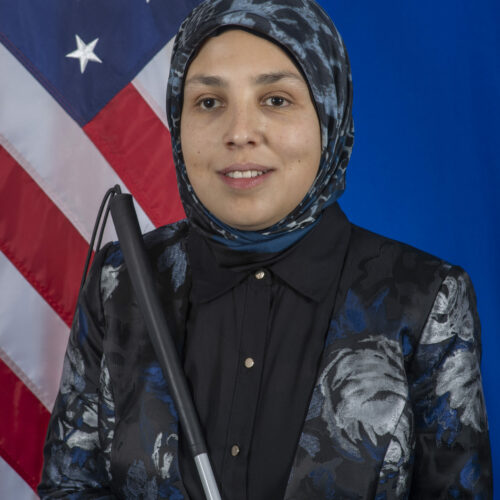 This is a photo of Sara Minkara. She wears a dark blue and black blazer with a patterned hijab. She holds a cane and stands in front of an American flag. She is smiling.