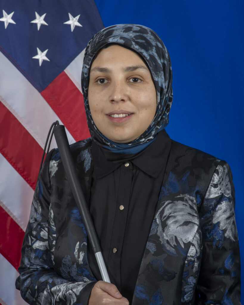 This is a photo of Sara Minkara. She wears a dark blue and black blazer with a patterned hijab. She holds a cane and stands in front of an American flag. She is smiling.