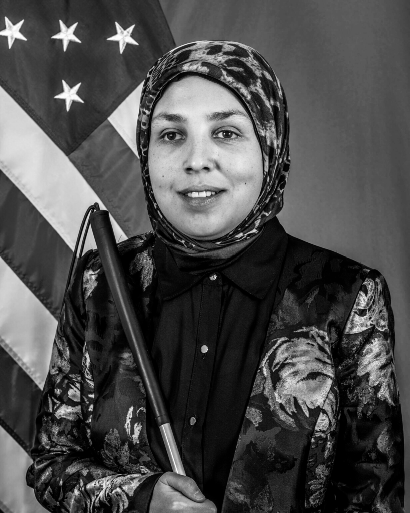 This is a black and white photo of Sara Minkara. She is posing for this photo in front of an American flag. She has a cane in hand.