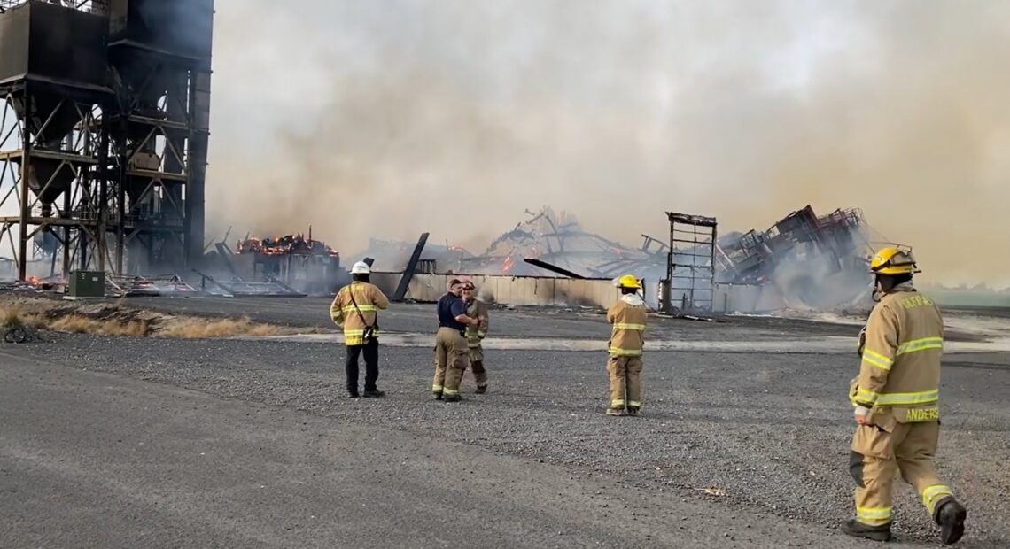 Firefighters stand near a pile of gray rubble as smoke billows upward.