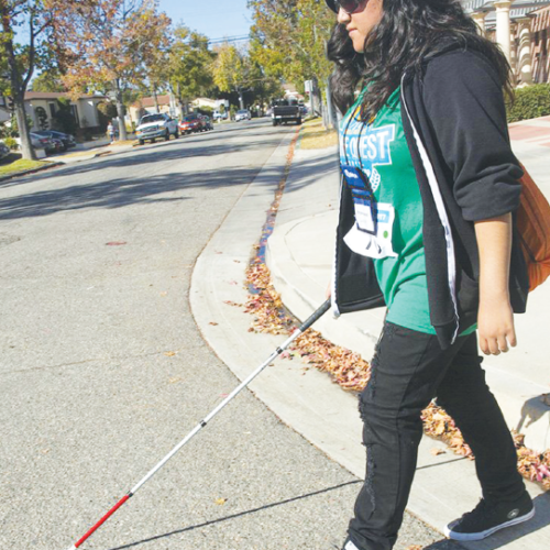 young teen with a visual impairment walks down a street with a cane.