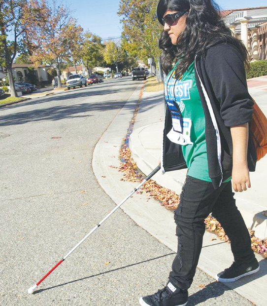 young teen with a visual impairment walks down a street with a cane.