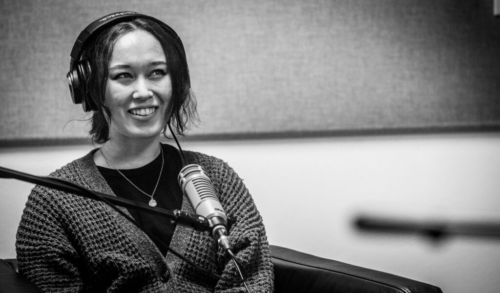 Photo of Zoe Hana Mikuta with headphones on in front of a microphone. She smiles and looks up.