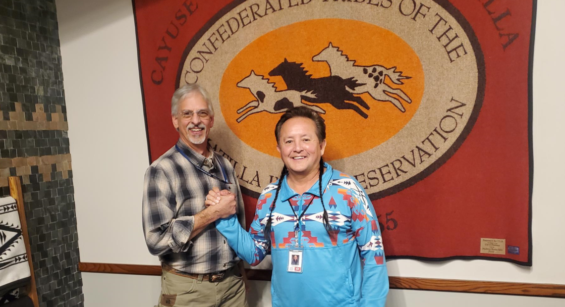 Two men stand in front of the CTUIR flag clasping hands and smiling toward the camera. The man on the left is white, tall, wearing a plaid shirt and khaki pants, the man on the right is shorter, Native, his hair braided into two braids, pulled over his shoulder, he's wearing a blue patterned shirts with geometric, patterns and jeans.