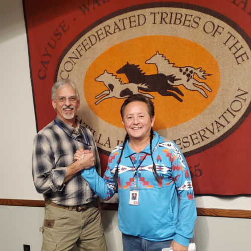 Two men stand in front of the CTUIR flag clasping hands and smiling toward the camera. The man on the left is white, tall, wearing a plaid shirt and khaki pants, the man on the right is shorter, Native, his hair braided into two braids, pulled over his shoulder, he's wearing a blue patterned shirts with geometric, patterns and jeans.