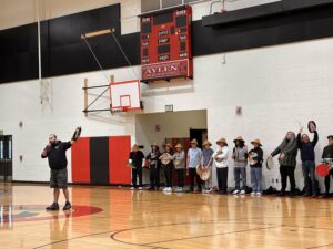 A man in a black sweatshirt stands in the middle of a gymnasium with a microphone. A row of people stand against the wall behind him.