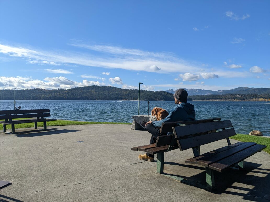 A man in jeans with a gray beanie sits on a bench with his cocker spaniel looking out at the blue water and dark blue mountains ahead.