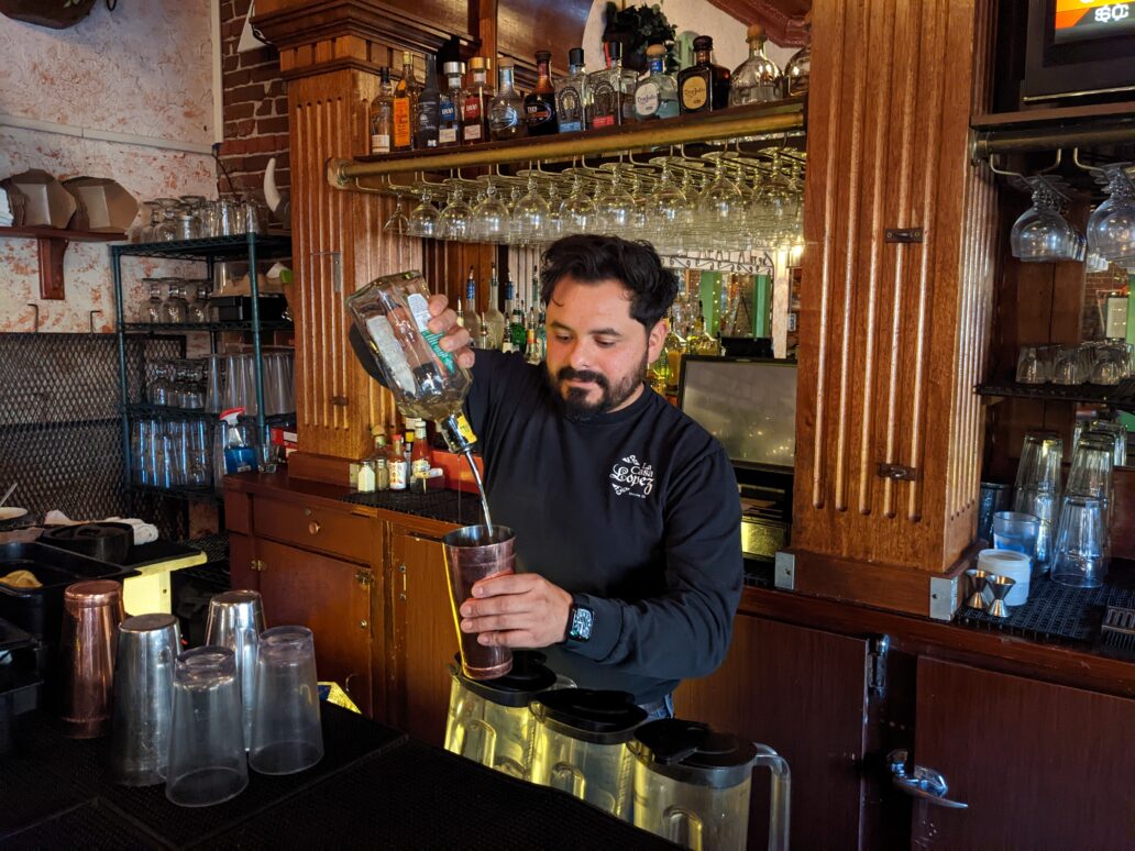 A man with dark hair pours a drink into a copper cup as he stands in front of a wooden bar with glassware hanging upside down behind him.