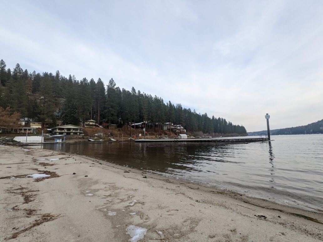 A beach with patches of snow is shown against dark green trees near Lake Coeur d'Alene. 