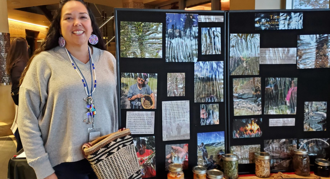 Teara Farrow Ferman stands in front of a display of photographs and containers documenting and displaying first foods. Farrow Ferman wears large, colorful, circular beaded earrings, she has long, dark hair and is smiling.