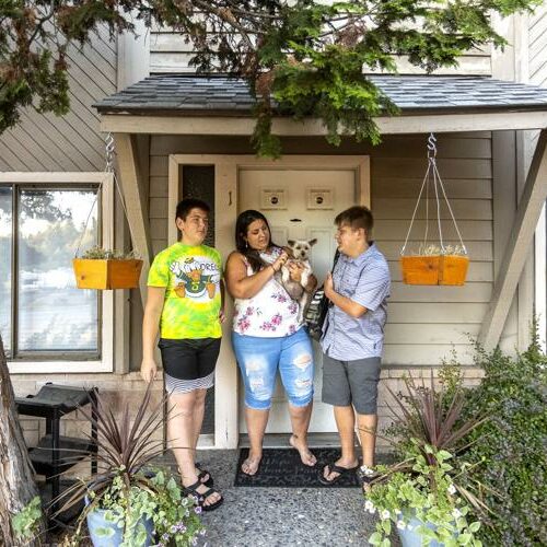 A mom and two sones stand in front of their rental home. She is holding a small dog.